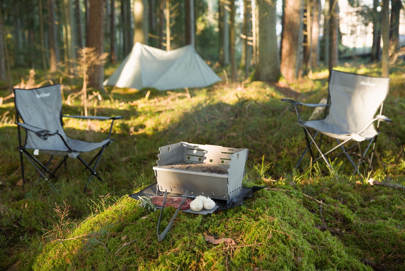 Titan1 titanium fire pit on moss between two camp chairs, tent pitched behind