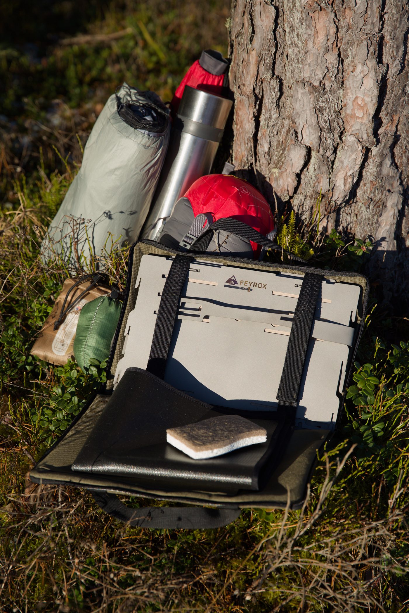 Titan1 packed flat against a pine trunk beside a thermos and rolled base mat