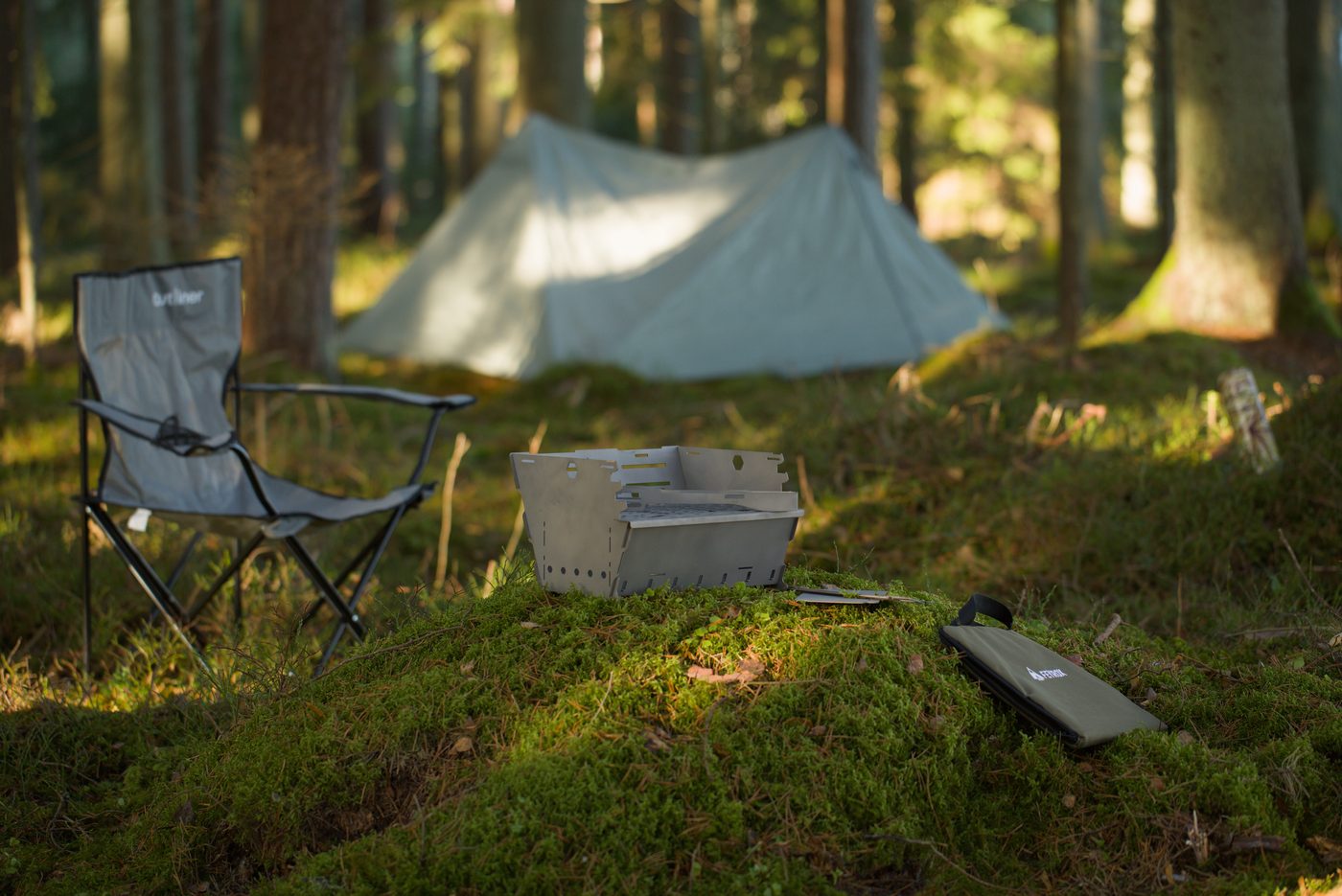 Titan1 titanium fire pit set up on moss beside a camp chair and tent, carry bag rolled alongside